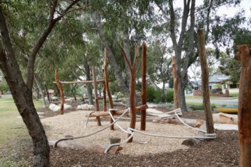 A nature playground with wooden climbing structures and ropes set among tall trees, surrounded by wood chips and grassy areas, with houses visible in the background.