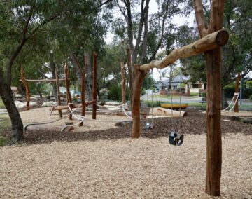 A natural playground with wooden climbing structures, swings, rope bridges, and woodchip ground cover, surrounded by trees and a residential street in the background.