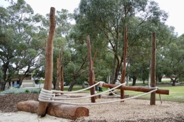 A natural playground featuring tall wooden poles and thick ropes for climbing, set on mulch and sand, surrounded by trees and grassy parkland.