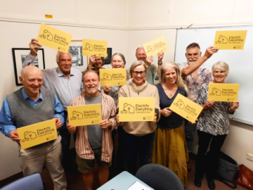 Nine smiling adults stand together indoors, holding yellow signs that read Electrify Everything coalition with a simple graphic of an electric plug. They appear to be participating in an advocacy or awareness event.