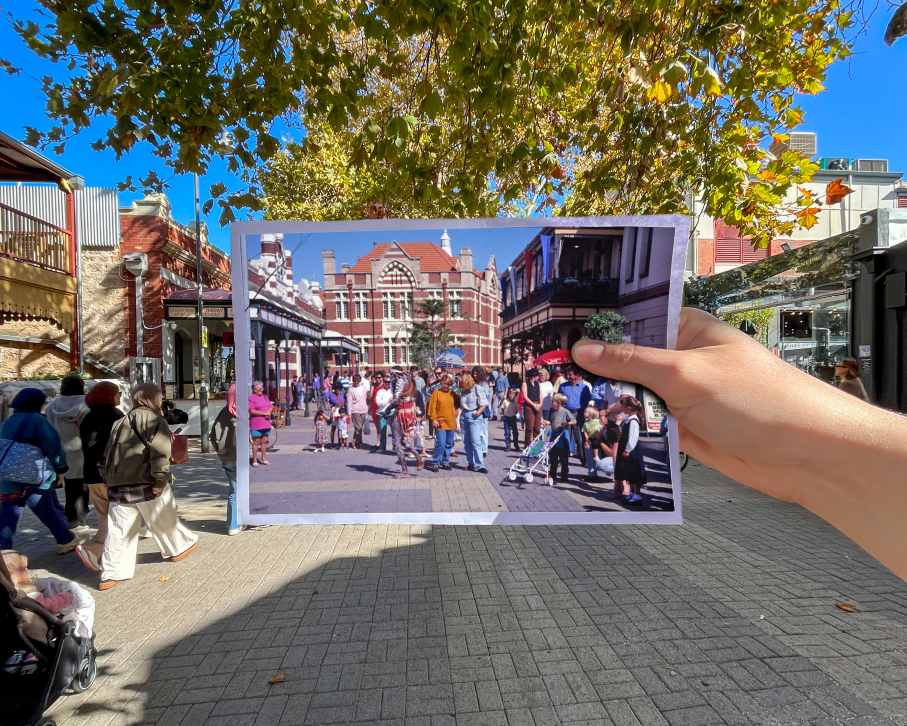 A hand holds an old photo of a busy street scene, aligning it with the same location in the present, which is now quieter, showing changes over time with similar buildings and trees in the background.
