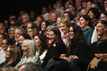 A large group of people seated closely together in an auditorium, smiling and laughing as they watch an event or performance. The audience includes adults of various ages and appearances.