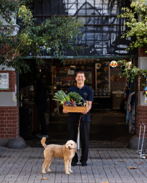 A smiling man stands outside a market entrance holding a crate of fresh vegetables, with a brown, curly-haired dog on a leash in front of him. Greenery and building details surround the arched entryway.