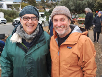 Two men wearing winter jackets, beanies, and scarves smile at the camera while standing outdoors at a community event. Other people and trees can be seen in the background on a cloudy day.