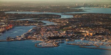Aerial view of a coastal city with marinas, docks, and numerous boats. Urban areas with buildings and homes stretch inland, with waterways and distant city skyline visible under a hazy sky.