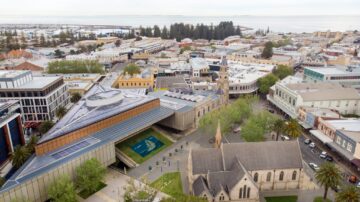 Aerial view of a coastal city with modern and historic buildings, including a triangular glass-roofed structure, a stone church, tree-lined streets, and the ocean in the background.