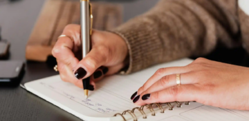 A person with dark nail polish writes in a spiral notebook with a pen. Their hand rests on the notebook, and they wear a simple ring. A phone and a brown case are partially visible on the dark table.