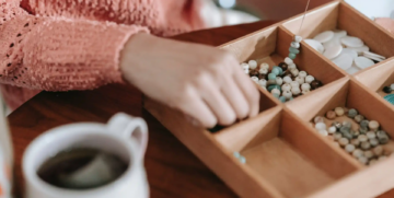 A person wearing a textured pink sweater selects beads from a wooden organizer with six compartments. A mug of tea sits nearby on a wooden table.