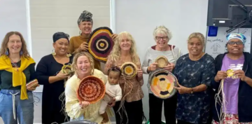A group of eight women and a baby stand together indoors, smiling and proudly holding handmade woven baskets of various sizes and colors. Basket-making materials are visible in some of their hands.