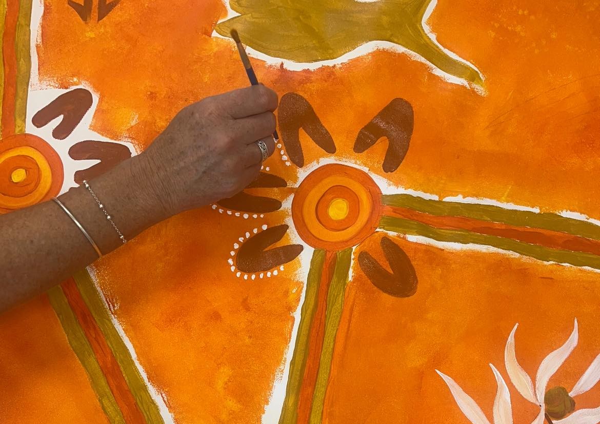 A hand holding a paintbrush adds white dots to a circular, abstract pattern on an orange canvas with earthy designs and lines.