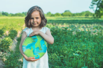 A young girl in a white dress stands in a green field, holding a cardboard cutout painted to look like the Earth, with trees and blue sky in the background.