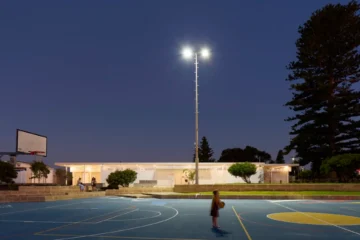 A child holding a basketball stands on an outdoor court at dusk, illuminated by bright overhead lights, with trees and buildings visible in the background.