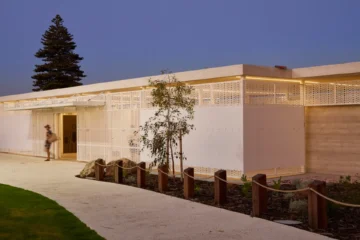A modern, single-story building with a white perforated facade and warm lighting at dusk; a person is walking towards the entrance along a curved pathway bordered by small trees and wooden posts.