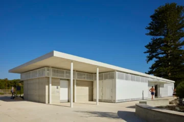 A modern public restroom building with clean lines, light-colored walls, and perforated white panels, set against a clear blue sky. Two people walk nearby on the concrete path, and a large tree stands at the right.