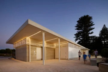 A modern public restroom building with concrete walls, white panels, and a flat roof, illuminated by warm lights at dusk. Several people, including children, walk near the entrance and along the walkway.