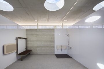 A spacious, modern accessible bathroom with high ceilings, overhead skylights, a fold-down shower seat, grab bars, a shower area, a wooden bench, and a baby changing station on the left wall.