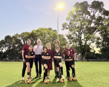 A group of four adults and four children in soccer uniforms stand on a grassy field with soccer balls, posing for a photo at sunset with trees and a tall light in the background.