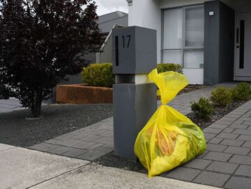 A yellow garbage bag sits on the pavement next to a modern mailbox labeled 17 outside a house with a gray tiled path and neatly trimmed bushes.