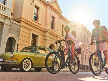 Two people wearing helmets ride bicycles past a parked vintage green car on a sunny day, with sunlight streaming over old, beige-colored buildings in the background.