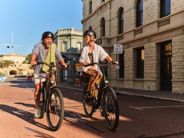 Two adults wearing helmets ride electric bikes on a sunny street lined with historic stone buildings. They are smiling and engaging in conversation, enjoying the pleasant day.