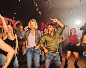 A group of people are dancing energetically in a lively, colorful club with string lights overhead. Two people are in the foreground, smiling and enjoying the upbeat atmosphere, surrounded by others also dancing enthusiastically.