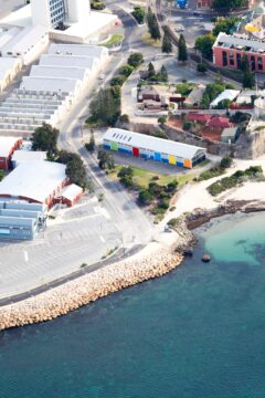 Aerial view of a coastal area with a rocky shoreline, clear water, parking lots, industrial buildings, colorful building, and houses surrounded by trees and streets.