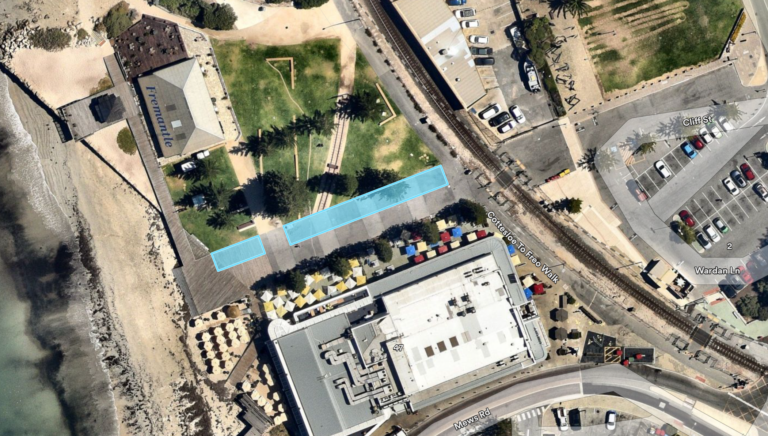 Aerial view of a coastal area with buildings, parking lots, beach, and green space. Blue rectangles highlight sections along a boardwalk between trees and a large building near the shoreline.