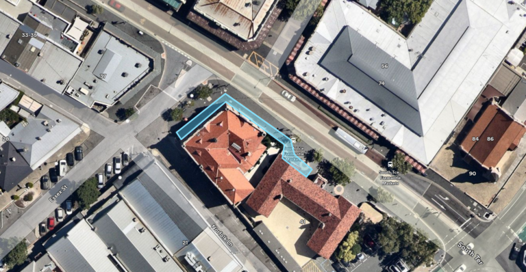 Aerial view of several buildings and streets; the main building in the center has a red roof and is outlined in blue, surrounded by roads, other structures, and a few parked cars.