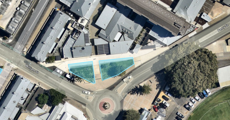 Aerial view of buildings and streets with a roundabout at the intersection of Pearl Street and Wilfred Street. Two irregular blue highlighted areas are shown near the roundabout, beside several trees and parked cars.