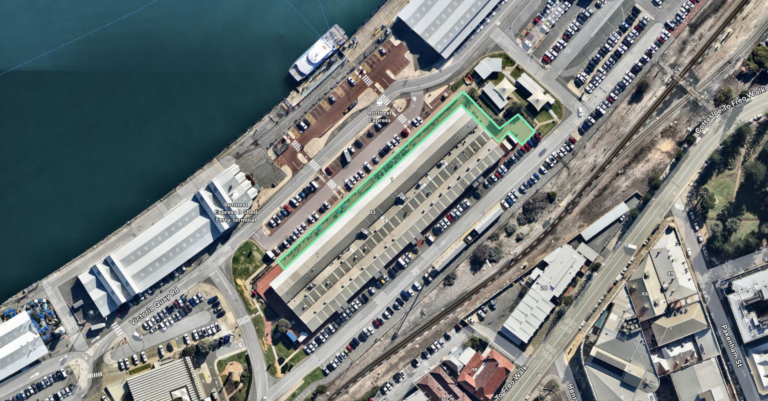 Aerial view of an industrial waterfront area with warehouses, a long building outlined in green, parked cars, nearby roads, and railway tracks.