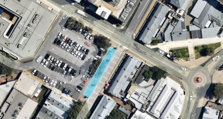 Aerial view of a city block with parking lots, buildings, streets, and a roundabout. A rectangular area in a parking lot is highlighted in blue next to Wellington St and Hillman St.