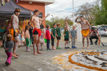 Indigenous performers in traditional attire and body paint dance before a group of children and adults on a paved area, with a colorful ground art design made of natural materials in the foreground.