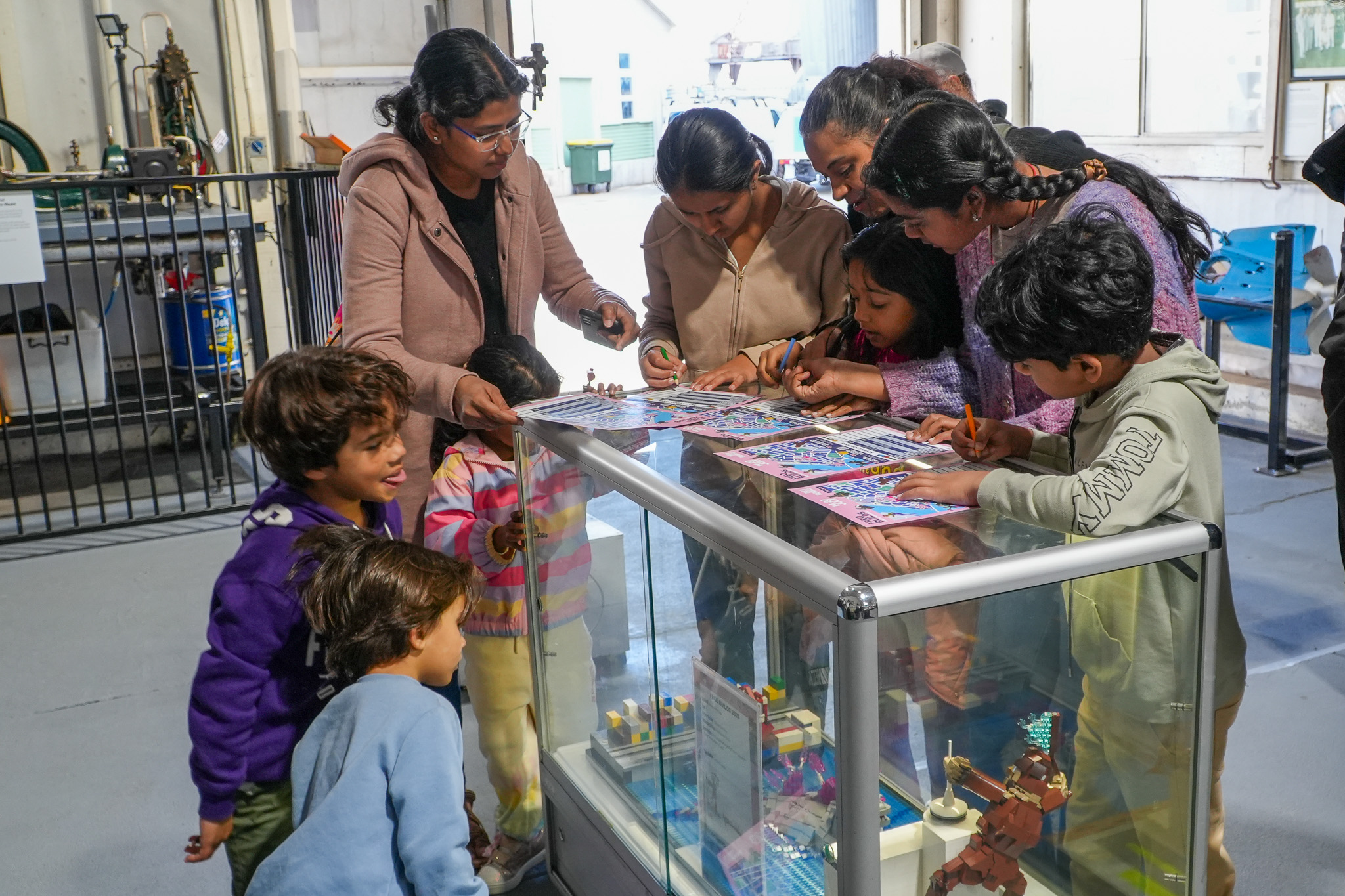A group of children and adults gather around a glass display case, coloring and drawing on paper, in a brightly lit indoor setting with educational or museum exhibits in the background.