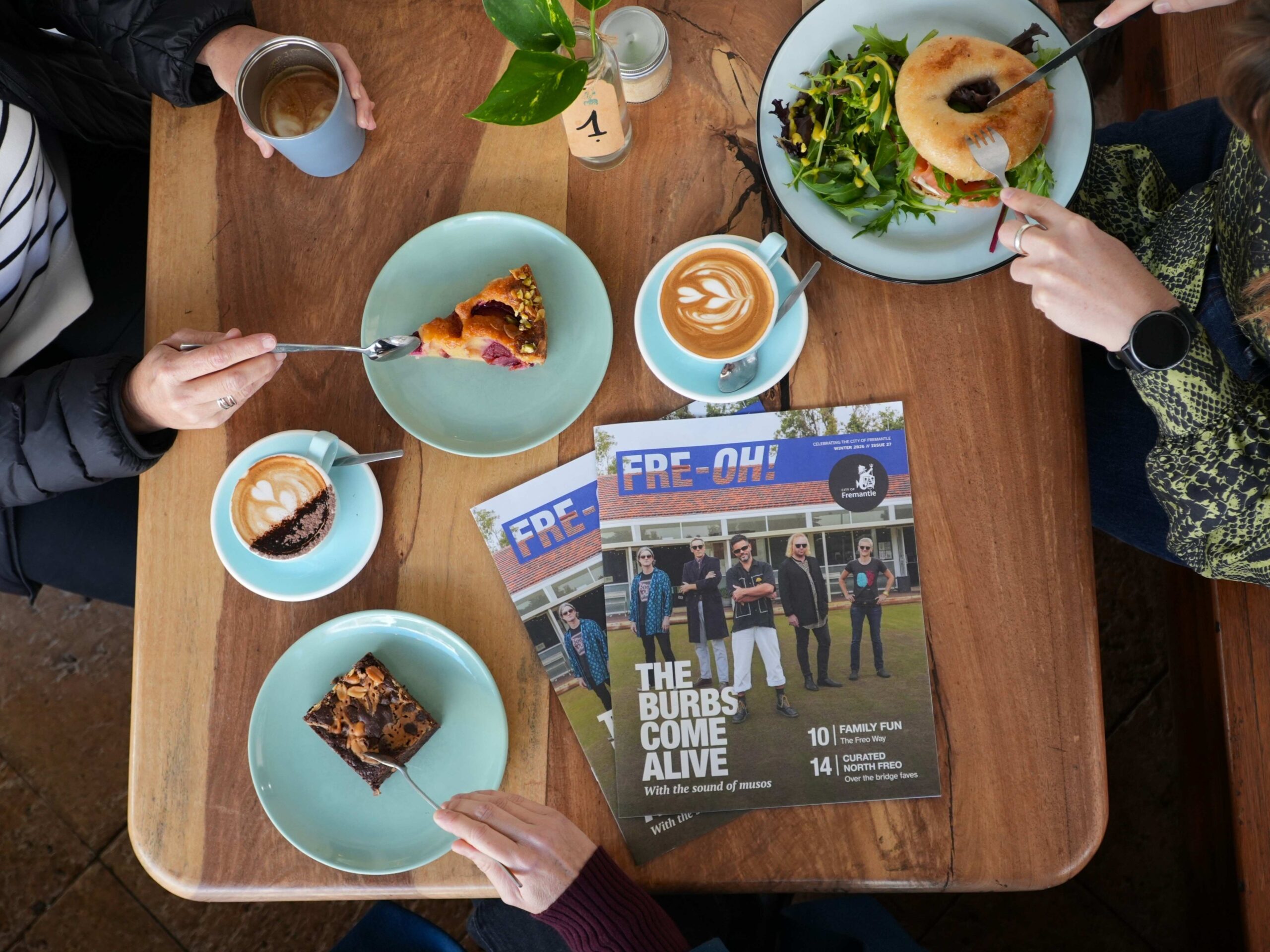 Three people sit at a wooden table with coffee, pastries, a bagel with salad, and a potted plant. Magazines featuring a band and the headline THE BURBS COME ALIVE are in the center of the table.