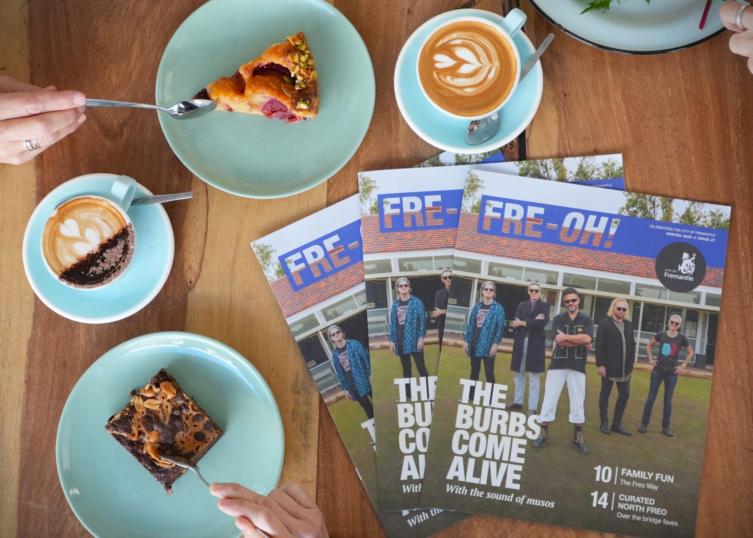 A table with three magazines, two cups of coffee with latte art, a chocolate brownie, a slice of fruit tart, and two hands—one holding a fork and the other holding a cup.