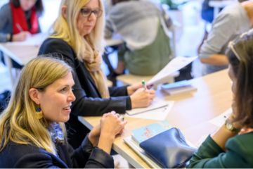 A group of women sit at a table engaged in conversation and writing on papers, with notebooks and a blue purse visible on the table in a bright, indoor setting.