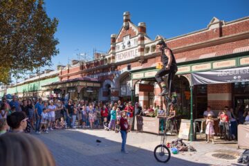 A street performer on a tall unicycle juggles clubs in front of a crowd outside Fremantle Markets. People, including children, watch the lively outdoor act on a sunny day. Historic brick building and shops are in the background.
