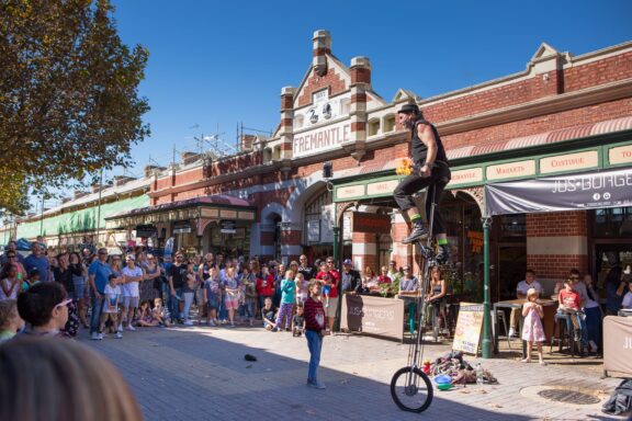 A street performer on a tall unicycle juggles clubs in front of a crowd outside Fremantle Markets. People, including children, watch the lively outdoor act on a sunny day. Historic brick building and shops are in the background.