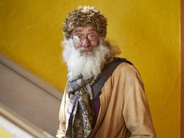 An elderly man with a long white beard and glasses stands indoors. He is wearing a shaggy brown hat, a tan coat, a patterned scarf, and a shoulder strap, with a bright yellow wall in the background.