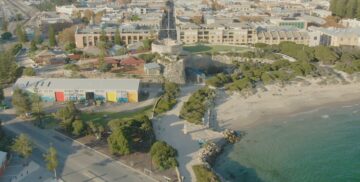 Aerial view of a coastal area with a sandy beach, green trees, pathways, colorful buildings, and a historic stone structure overlooking the water, with a town visible in the background.