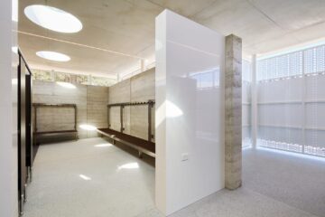 A modern locker room with concrete walls, long wooden benches, coat hooks, and natural light streaming through circular skylights and perforated privacy panels. The space appears clean and minimalistic.