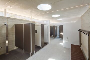 A modern public restroom with several toilet stalls, clean white walls, grey partitions, hooks on the wall, and round skylights in the concrete ceiling allowing natural light in.