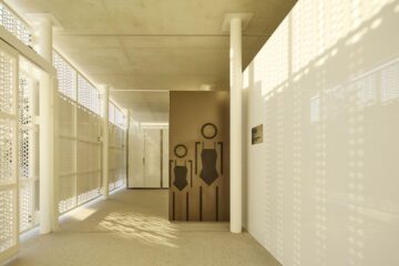A sunlit modern restroom entrance with minimalist geometric signs for men and women on a bronze panel, surrounded by perforated walls and white surfaces.