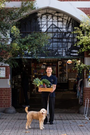 A person stands outside a market holding a box of fresh vegetables and herbs, smiling at the camera. A light brown, curly-haired dog on a leash stands beside them. Brick building and leafy trees frame the entrance.