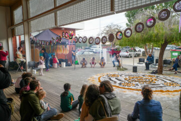 Children and adults watch a cultural performance outdoors. Several people sit on benches, while dancers perform in front of a large circular sand art. Artworks hang overhead and a tent is set up in the background near parked cars.