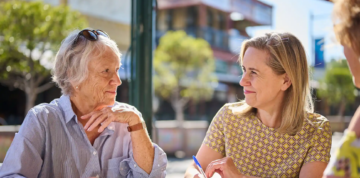 Two women, one older and one younger, sit at an outdoor table on a sunny day, smiling and talking. The younger woman holds a pen and paper. Trees and buildings are visible in the blurred background.
