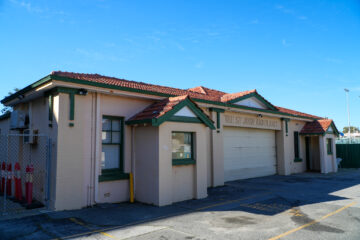 A single-story building with a red-tiled roof and cream walls, labeled The St. John Ambulance. It has green trim, a closed garage door, and is next to a chain-link fence with red bollards. The sky is clear and blue.