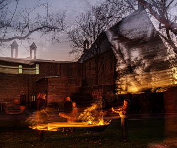 Four people stand around a fire pit at dusk on grass, with motion blur creating ghostly effects. Bare trees and an old brick building with chimneys are visible in the background.