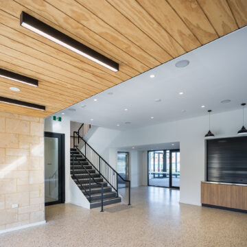 A modern, spacious interior with terrazzo flooring, a black metal staircase, wood panel ceiling, pendant lights, a stone feature wall, large windows, and a service counter. The space is brightly lit and minimalistic.