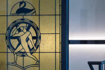 Stained glass window featuring an abstract figure pushing a large wheel, with geometric patterns and a swan-like design above, next to a modern, softly lit interior wall.